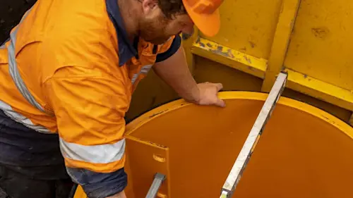 Construction worker in orange safety gear inspecting the interior of a orange industrial container.