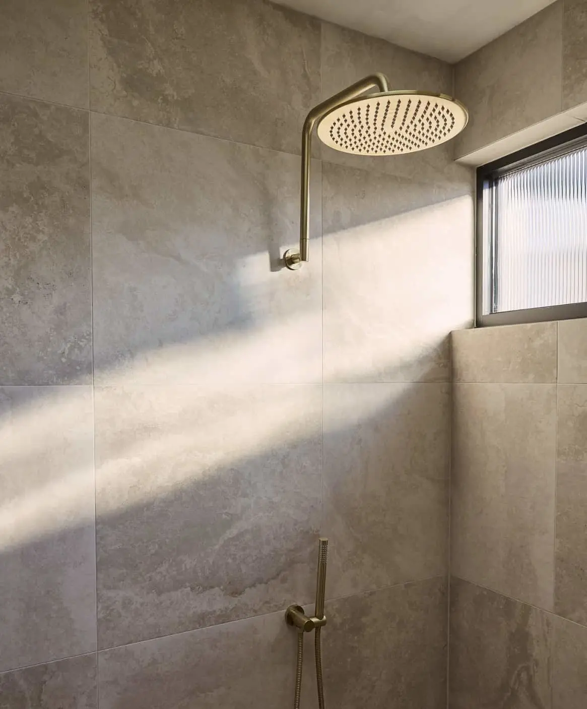Modern shower with brass rainfall showerhead and handheld sprayer in a stone-tiled bathroom with window.