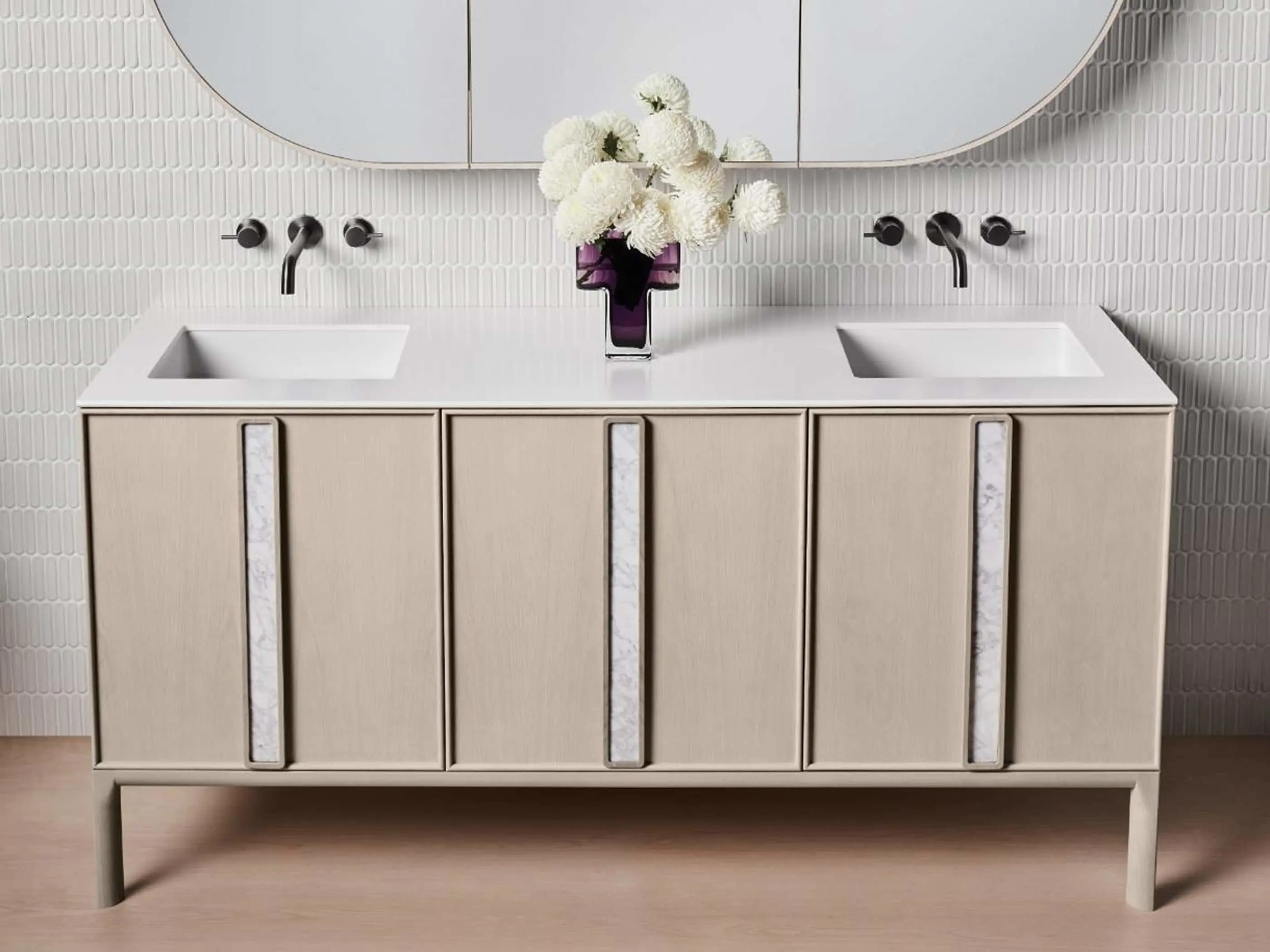 Modern bathroom vanity with dual sinks, light wood cabinets, white tile backsplash, and purple vase with white flowers.