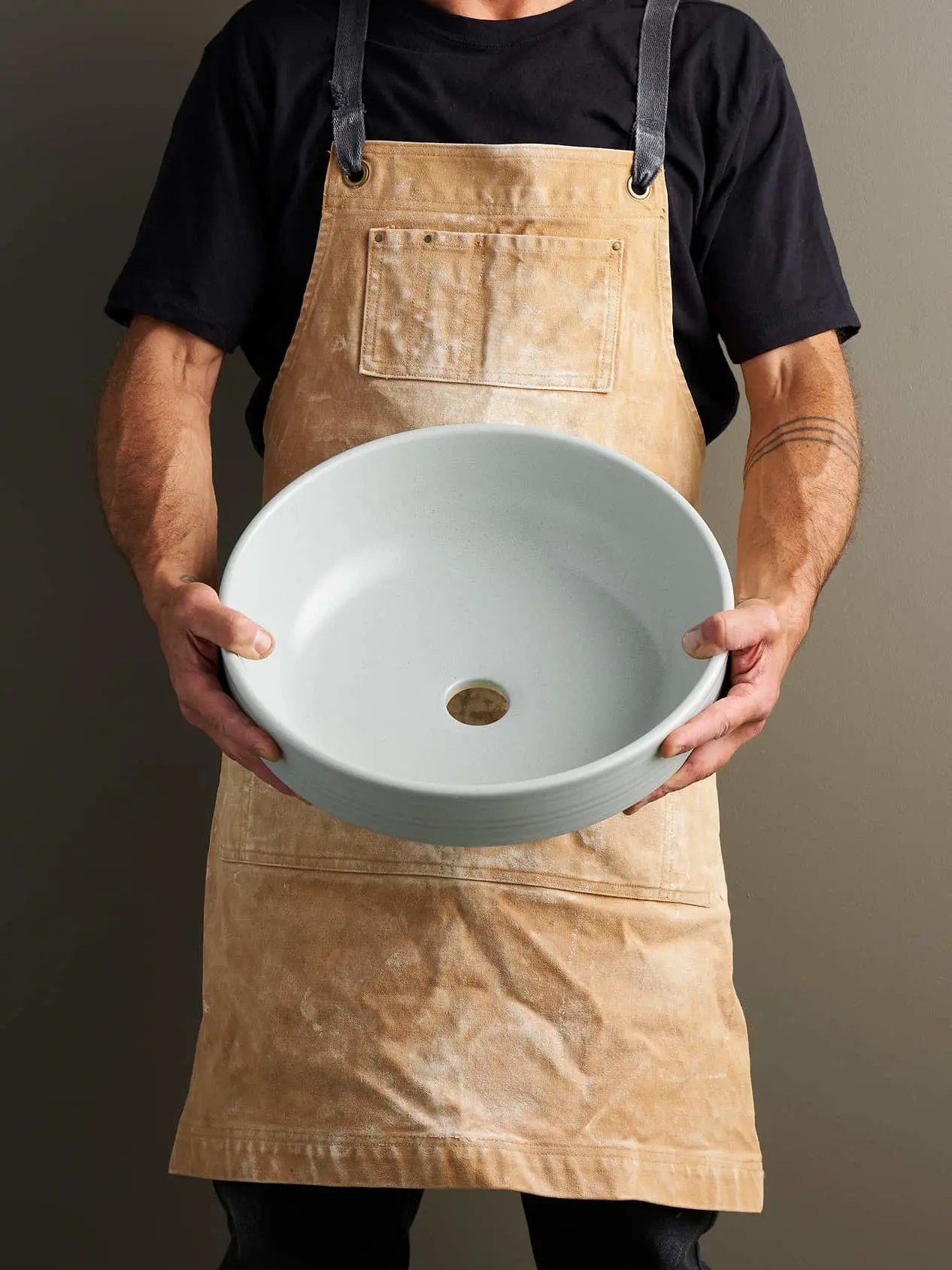 Person wearing tan canvas apron holding a white ceramic vessel sink with drain hole against neutral background.