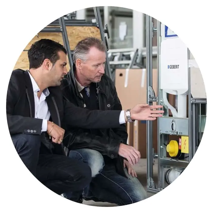 Two men in business attire examining a Geberit plumbing installation system with metal framework in a workshop setting.
