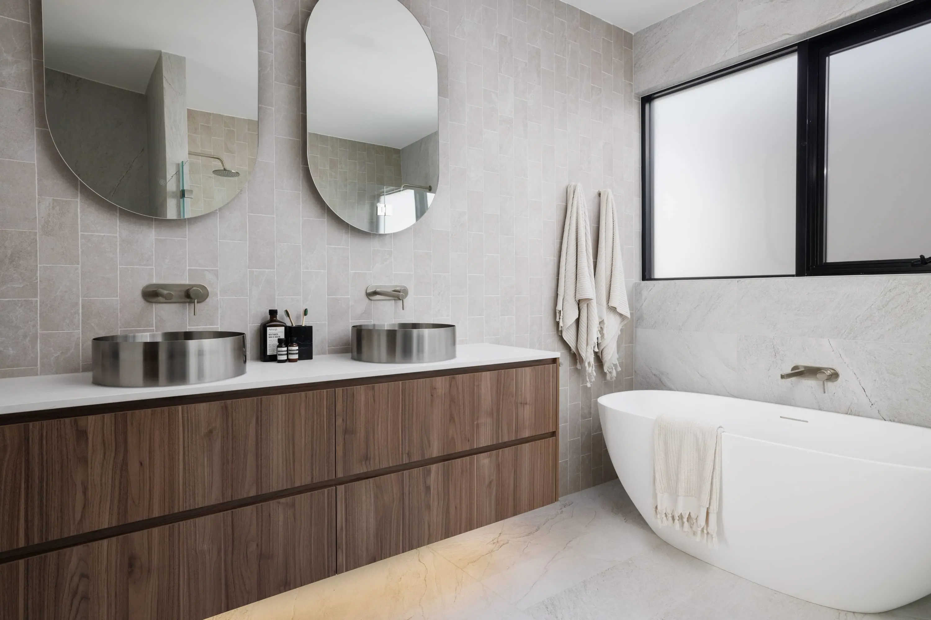 Modern bathroom with wooden vanity, dual metal sinks, round mirrors, beige tiled walls, and freestanding white bathtub.