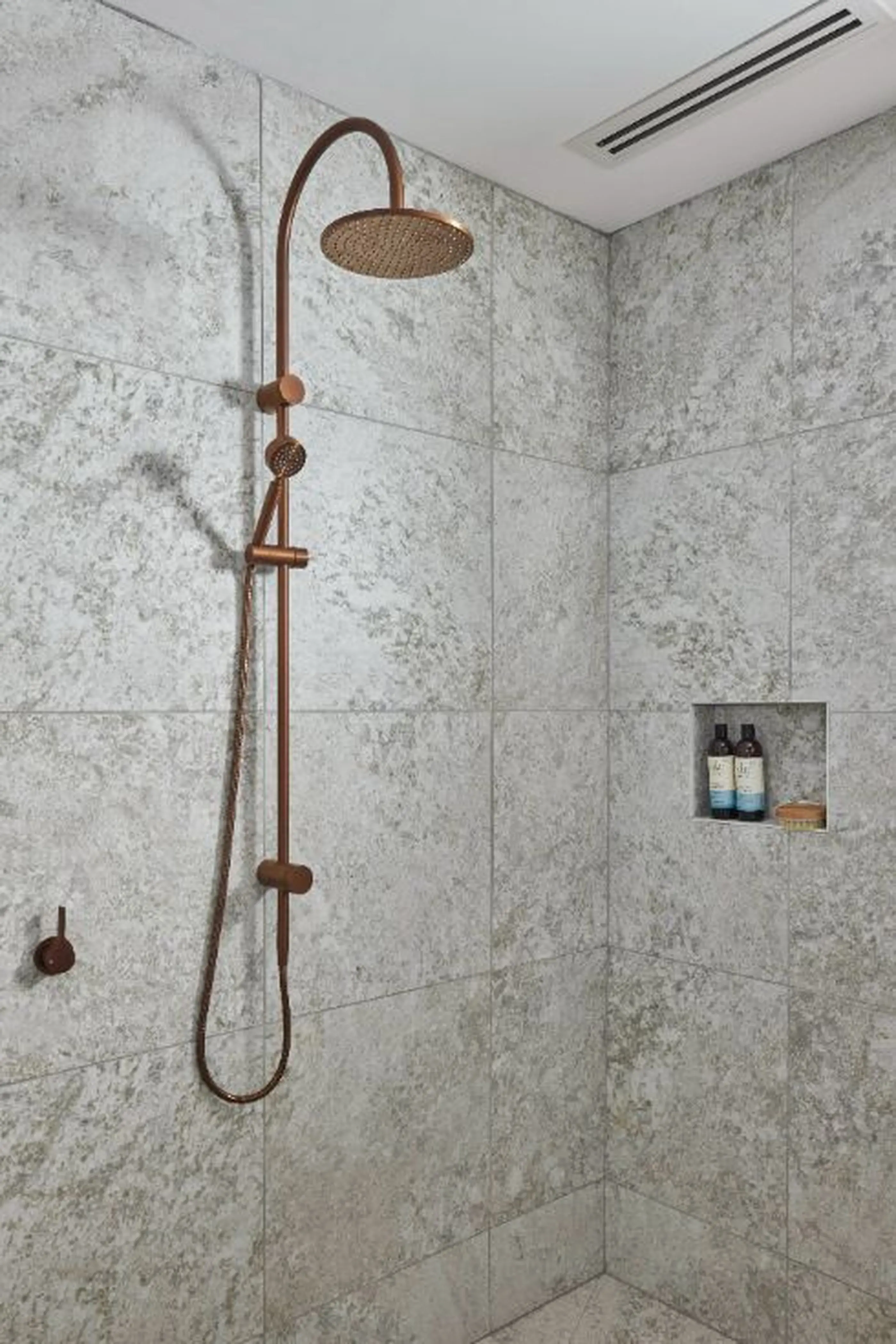 Modern shower with copper rainfall showerhead, marble-look tile walls, and built-in niche holding bath products.
