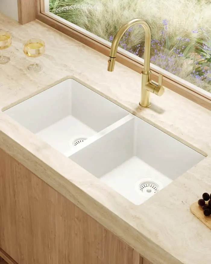 Modern white double kitchen sink with brass faucet on marble countertop, window overlooking purple wildflowers.