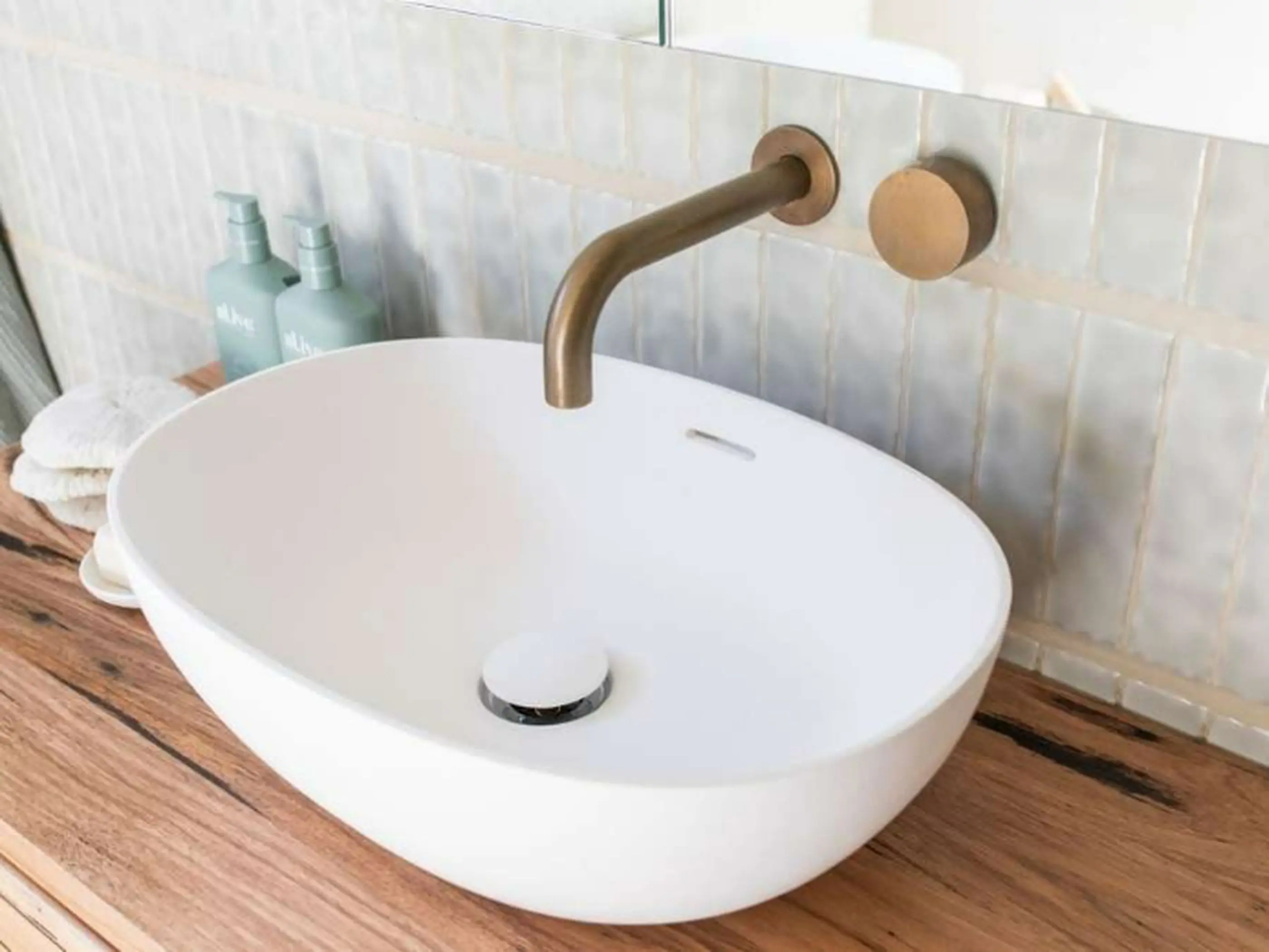 Modern white oval bathroom sink with brass wall-mounted faucet on wooden countertop with light tile backsplash.