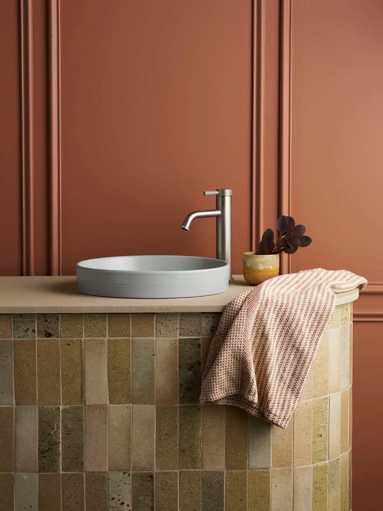 Modern bathroom sink with white vessel basin, brass faucet, and pink towel against terracotta walls and beige tile.