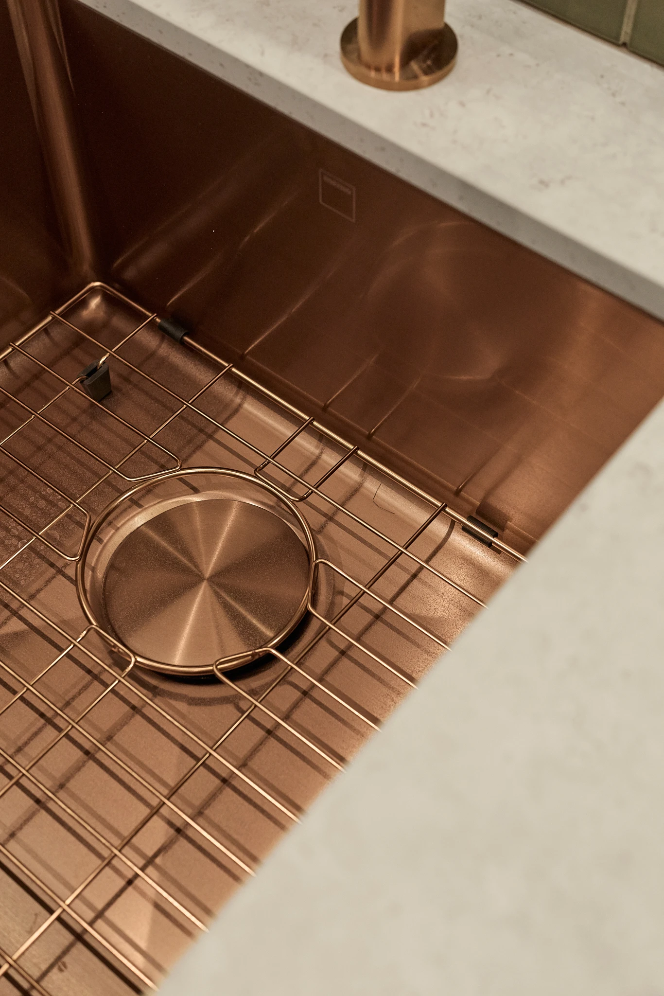 Copper kitchen sink with wire rack and drain cover, set in white countertop with matching copper faucet base visible.