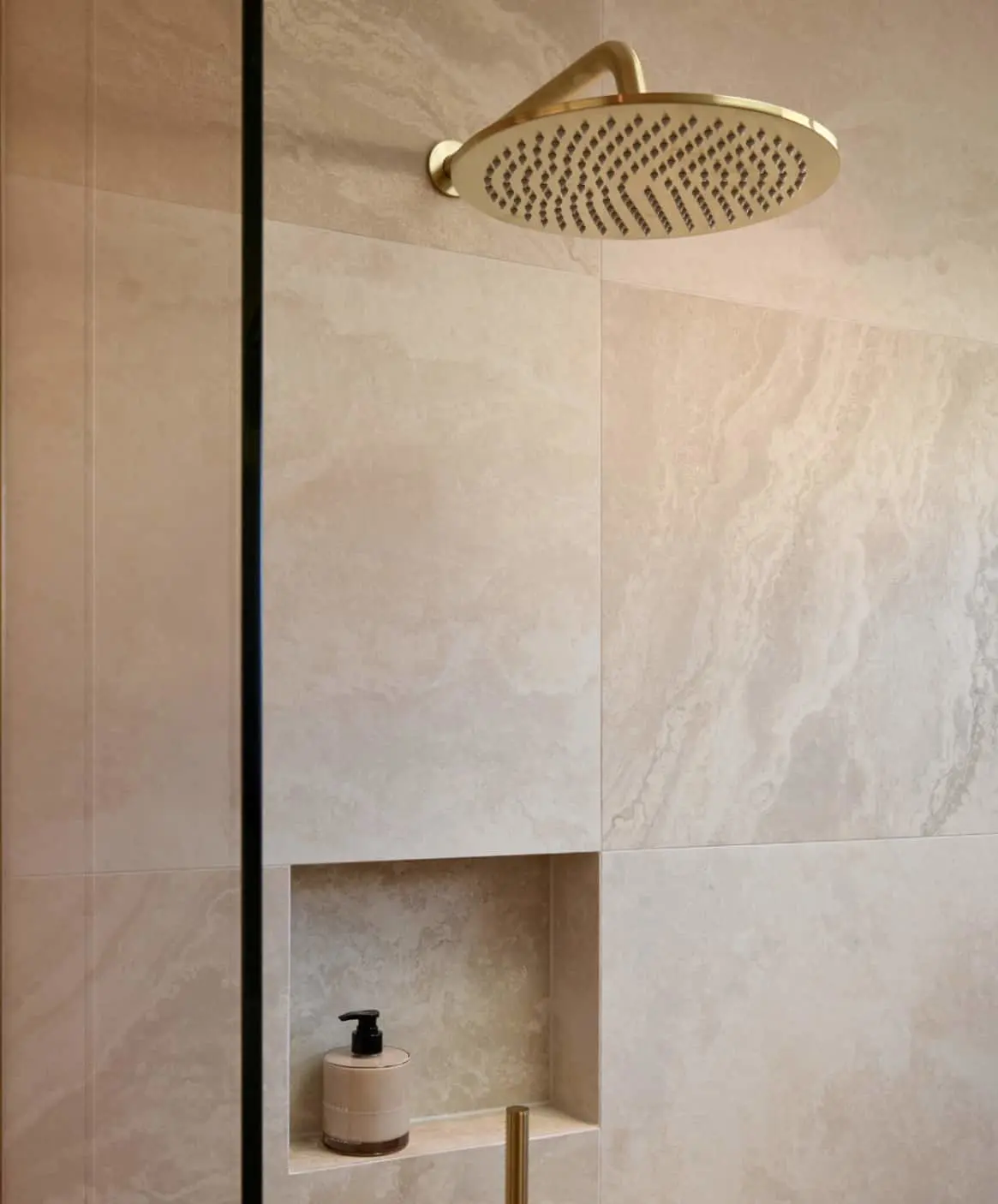 Modern bathroom with gold rainfall showerhead, brass fixtures, and beige soap dispenser in a marble-tiled shower niche.