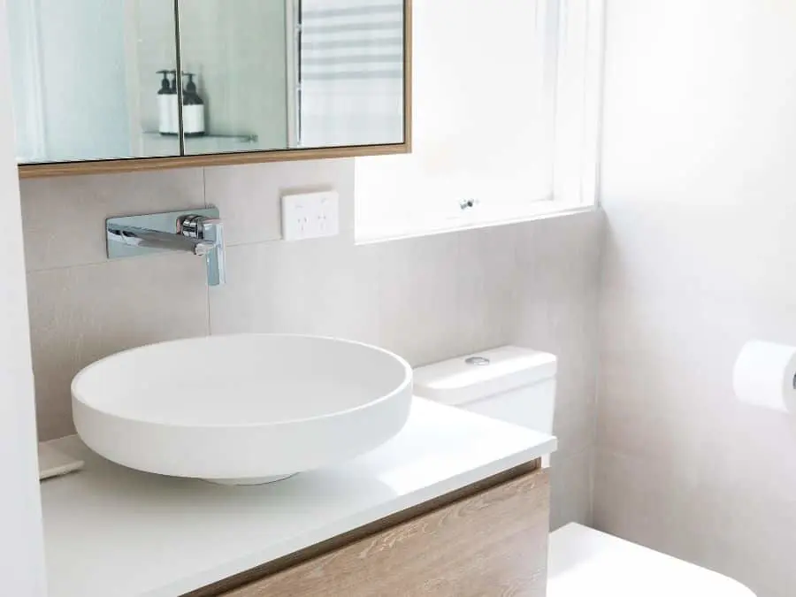 Modern bathroom with round white vessel sink, wooden vanity, wall-mounted faucet, and medicine cabinet mirror.