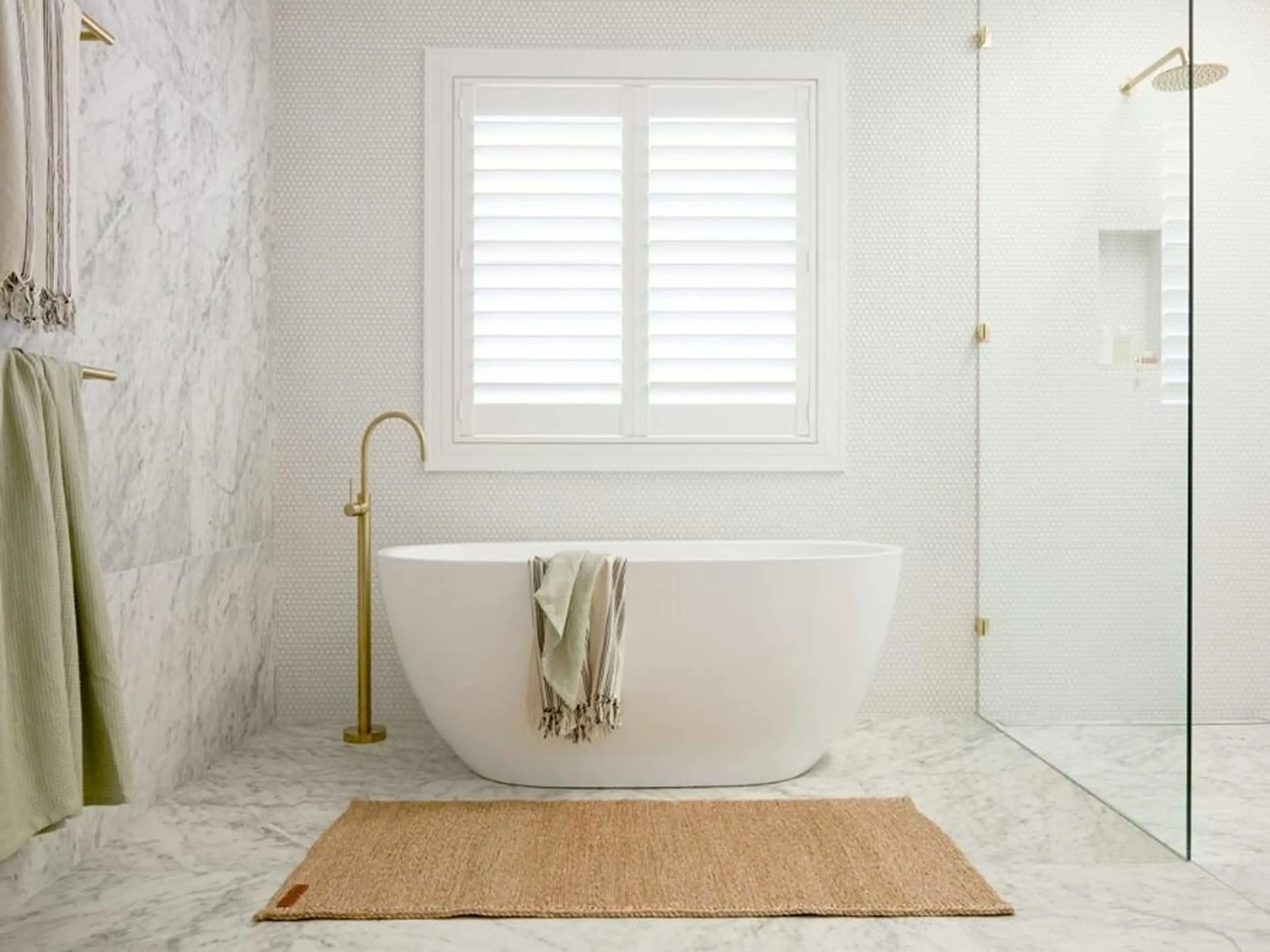 Modern white bathroom with freestanding tub, brass fixtures, marble walls, window with shutters, and jute rug.