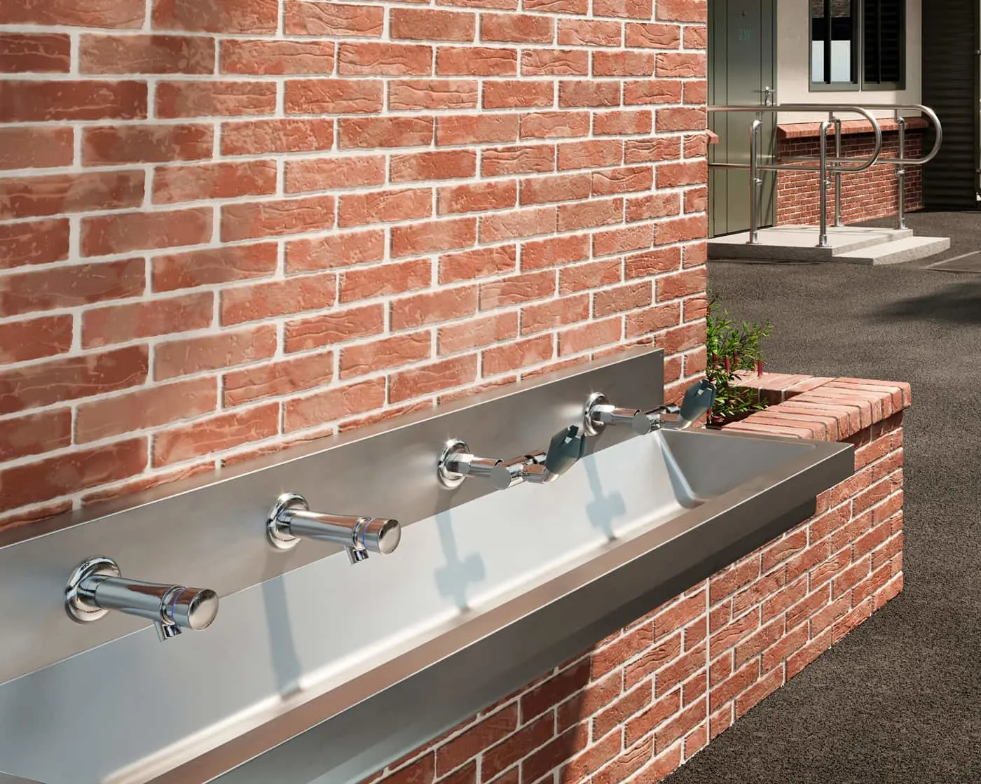Stainless steel outdoor handwashing station with multiple faucets mounted on red brick wall, with building entrance visible.