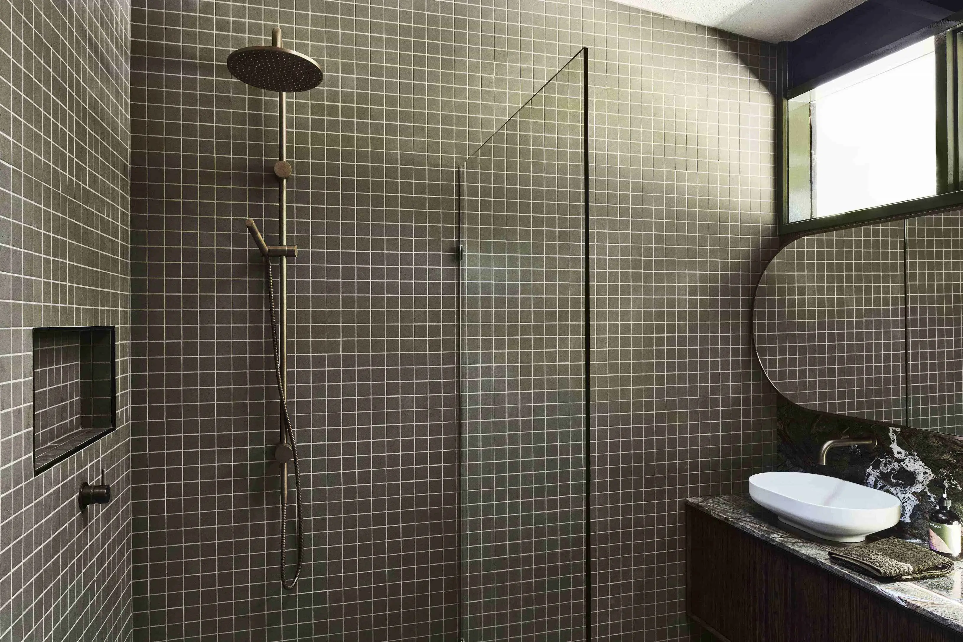 Bathroom with dark green mosaic tiles, brass shower fixtures, glass screen, marble vanity, white basin, and round mirror.