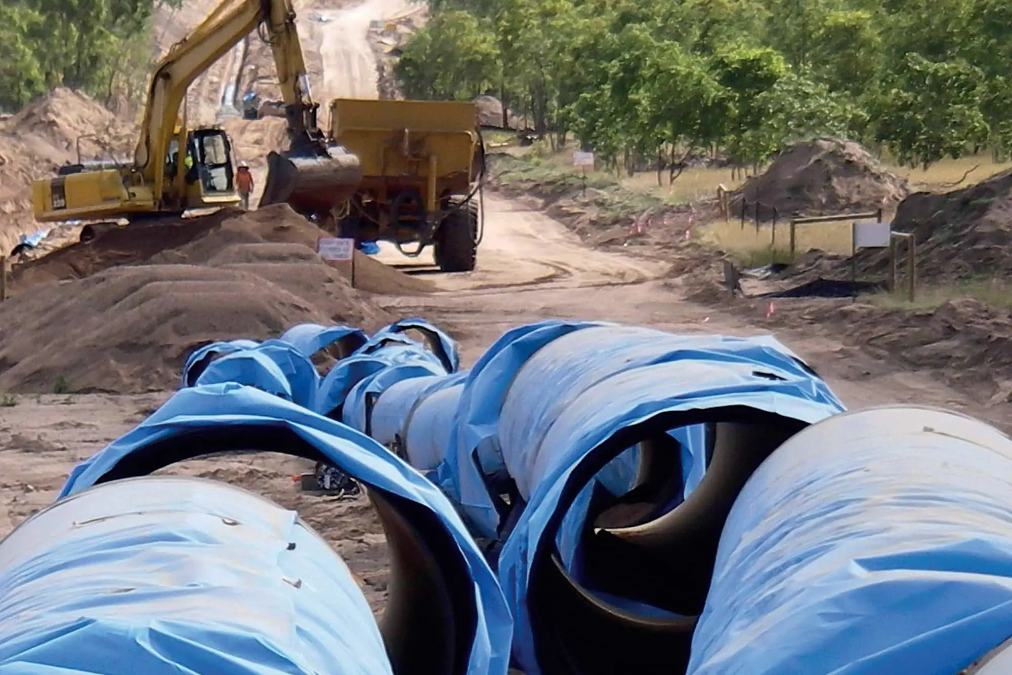 Blue water pipes at construction site with yellow excavator and dump truck working on dirt road surrounded by trees.