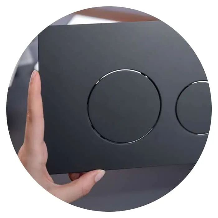 Hand holding a dark gray flush plate with two circular buttons for toilet flushing.