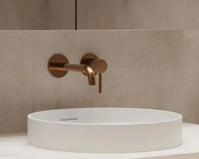Modern white circular basin with copper wall-mounted faucet against a beige textured wall in a minimalist bathroom.
