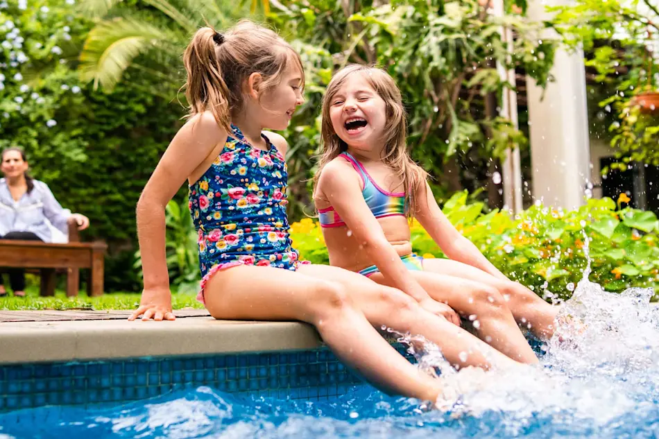 Two children in colorful swimsuits sitting at pool edge, laughing and splashing water in a lush garden setting.