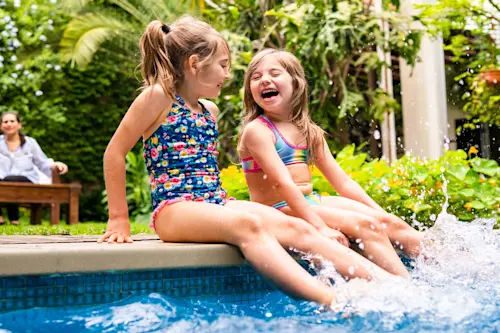 Two children in colorful swimsuits sitting at pool edge, laughing and splashing water in a lush garden setting.