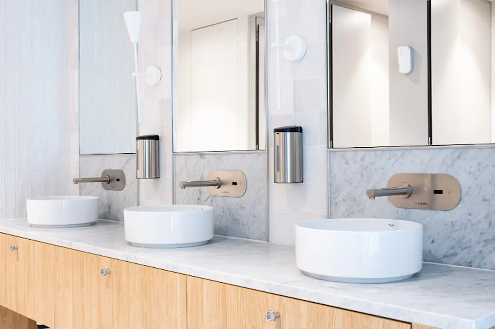 Modern bathroom with three white vessel sinks, wall-mounted faucets, marble backsplash, and wooden vanity cabinets.