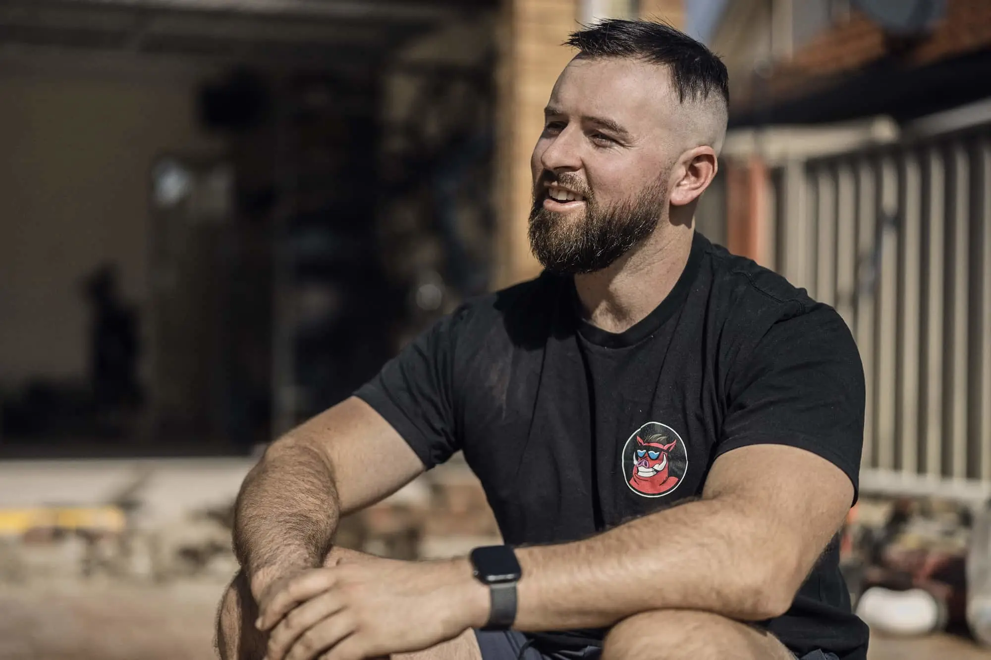A bearded man in a black graphic t-shirt and smartwatch sits outdoors, smiling and looking to the side.