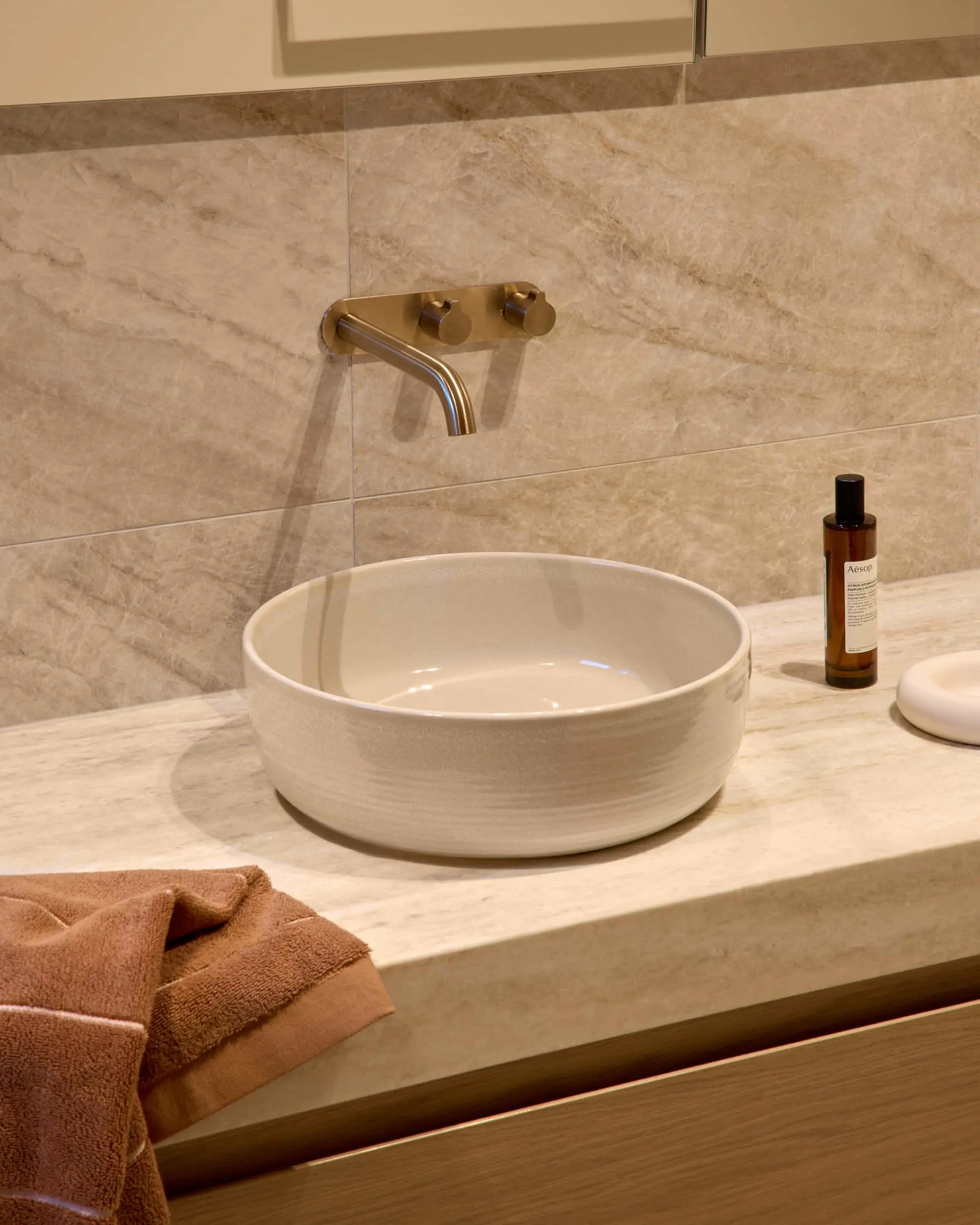 White round vessel sink on marble countertop with brass wall-mounted faucet and beige tiled wall.