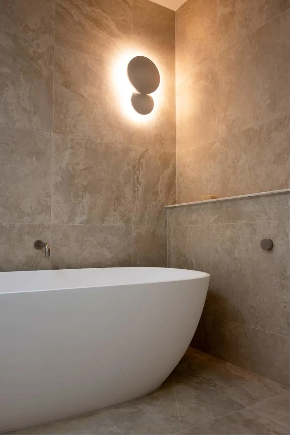 Modern bathroom with white freestanding tub, beige stone walls, and circular backlit wall sconce in corner.