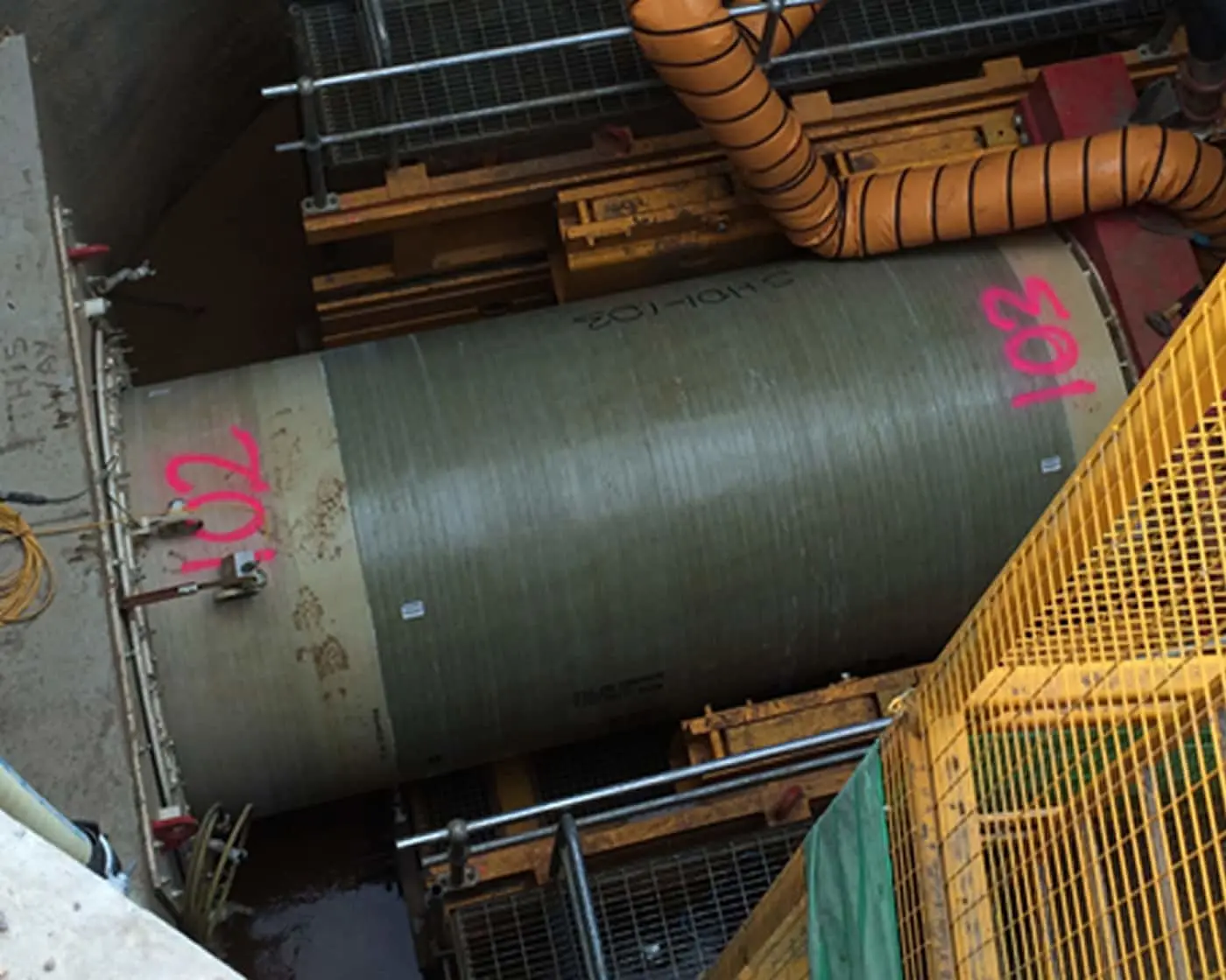 Large industrial metal cylinder with pink markings in a construction site, surrounded by orange ventilation ducts and safety railings.