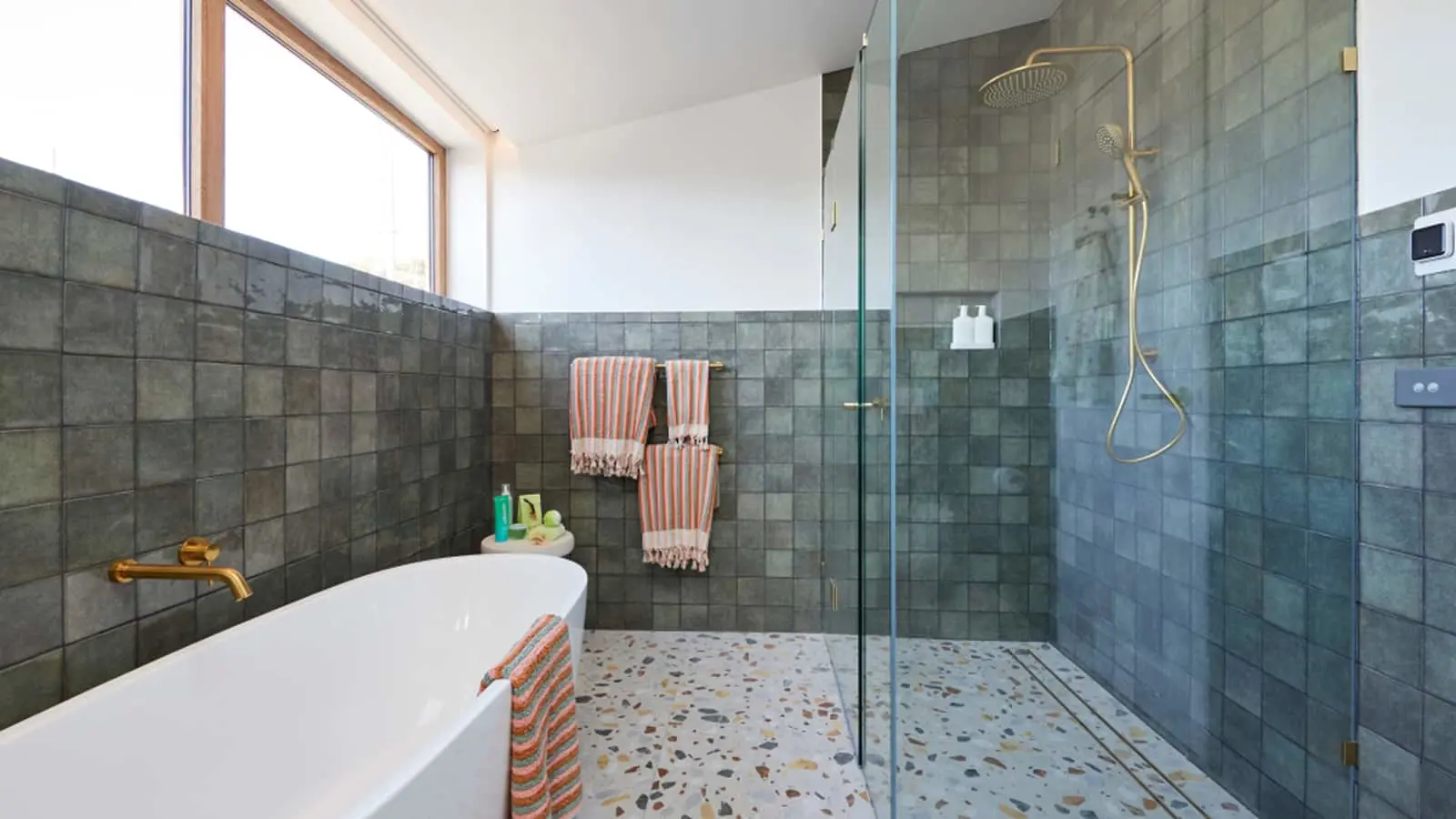 Modern bathroom with gray tile walls, white bathtub, glass shower enclosure, and orange striped towels on terrazzo floor.