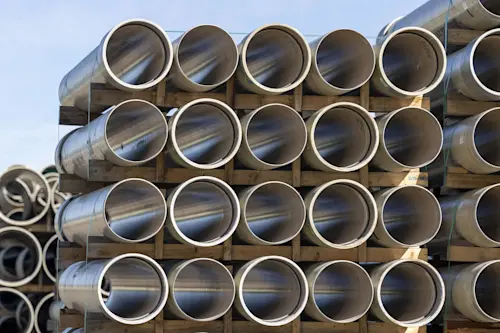Stacked metal pipes with threaded ends arranged in rows on wooden supports against a blue sky background.