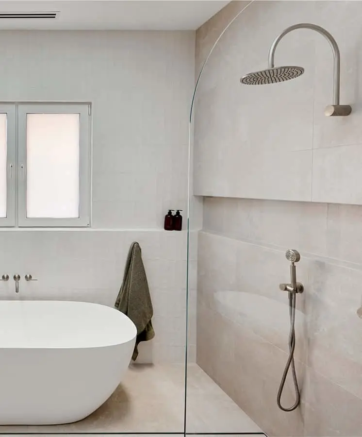 Modern minimalist bathroom with white freestanding tub, glass shower partition, and brushed metal rainfall showerhead.
