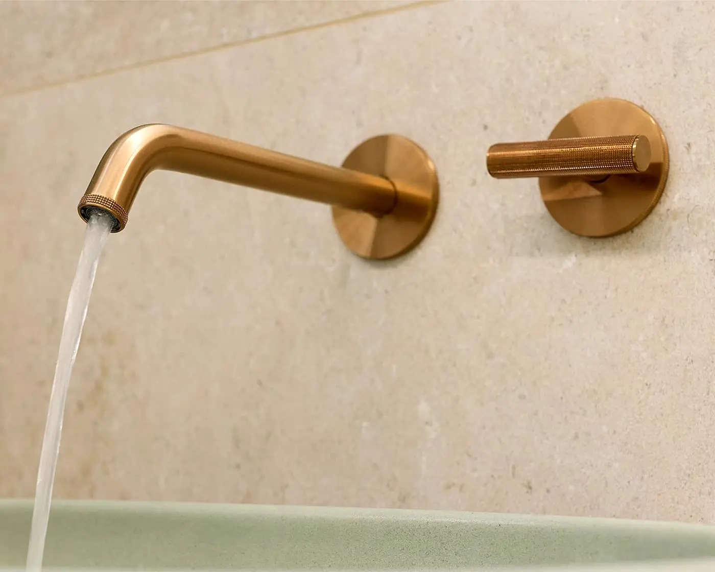 Brushed bronze wall-mounted bathroom faucet with water flowing into a light green basin against a beige stone wall.