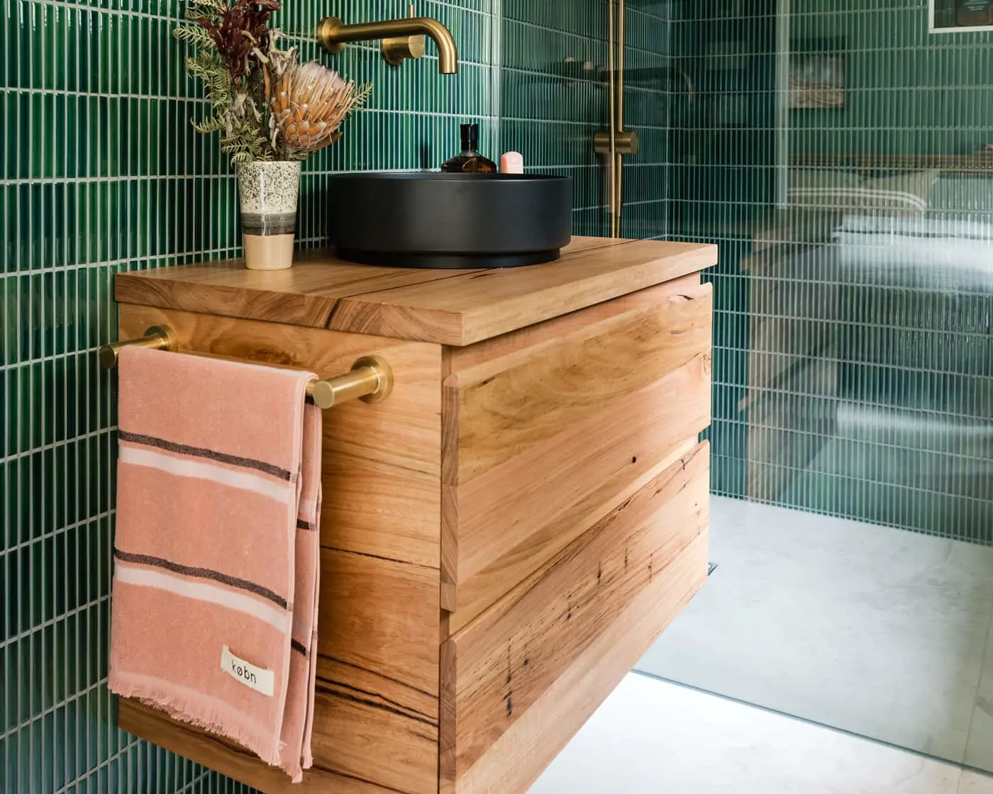 Modern bathroom with wooden vanity, black vessel sink, brass fixtures, green tile walls, and pink towel hanging on side.