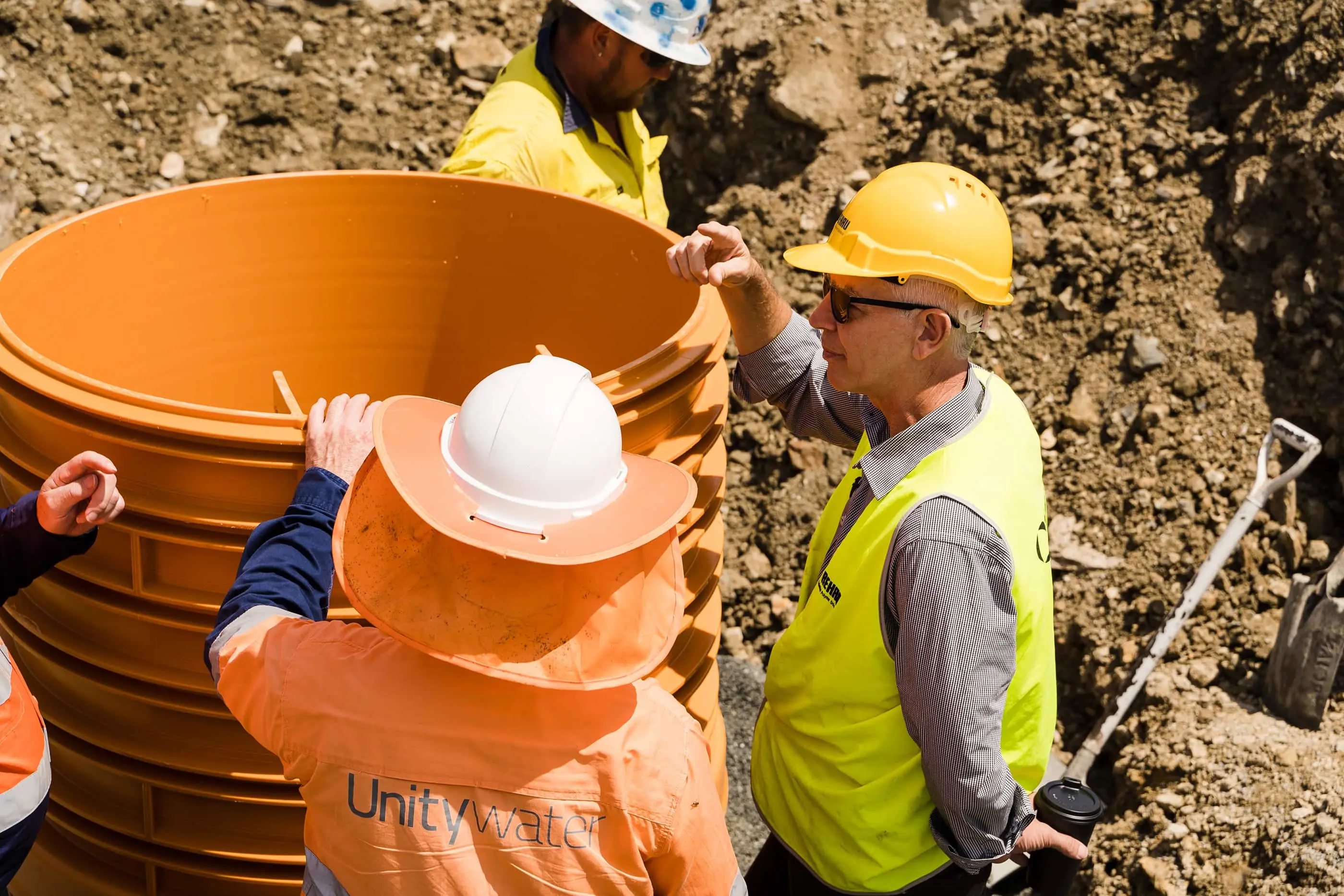 Construction workers in safety gear installing large orange water pipes at an excavation site.