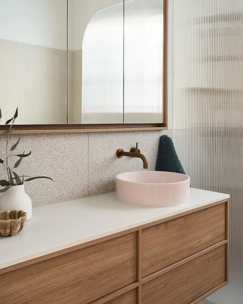 Modern bathroom with pink vessel sink, brass faucet, wooden vanity, mirrored cabinet, and textured glass partition.
