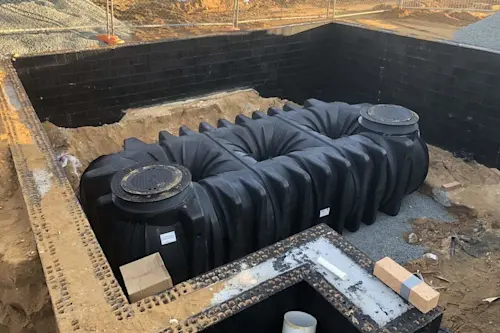 Large black underground water storage tank being installed in an excavated foundation with waterproofed walls.