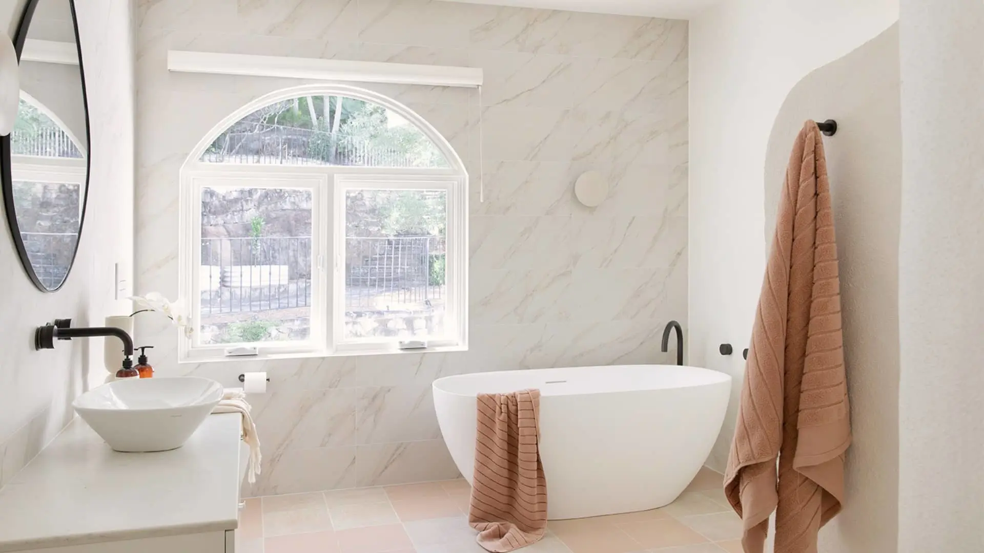 Modern bathroom with white marble walls, freestanding tub, vessel sink, arched window, and peach-colored towels.
