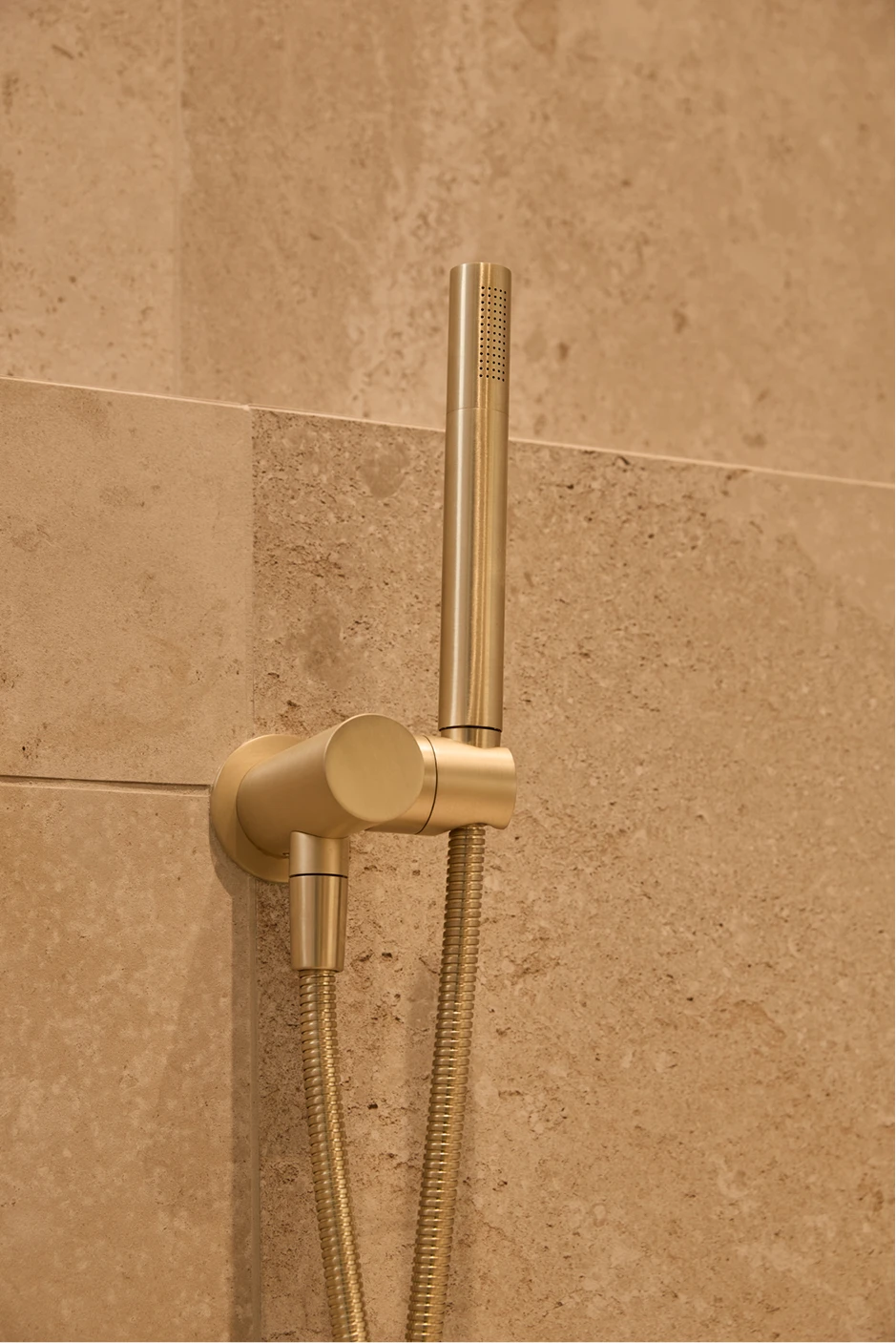 Gold handheld shower head with hose mounted on beige tiled bathroom wall.