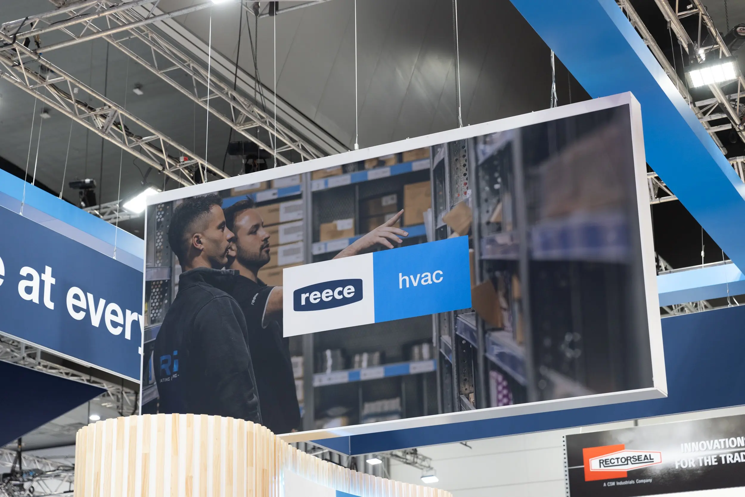 A large suspended display screen at a trade show showing the Reece HVAC logo and two workers looking at shelving.