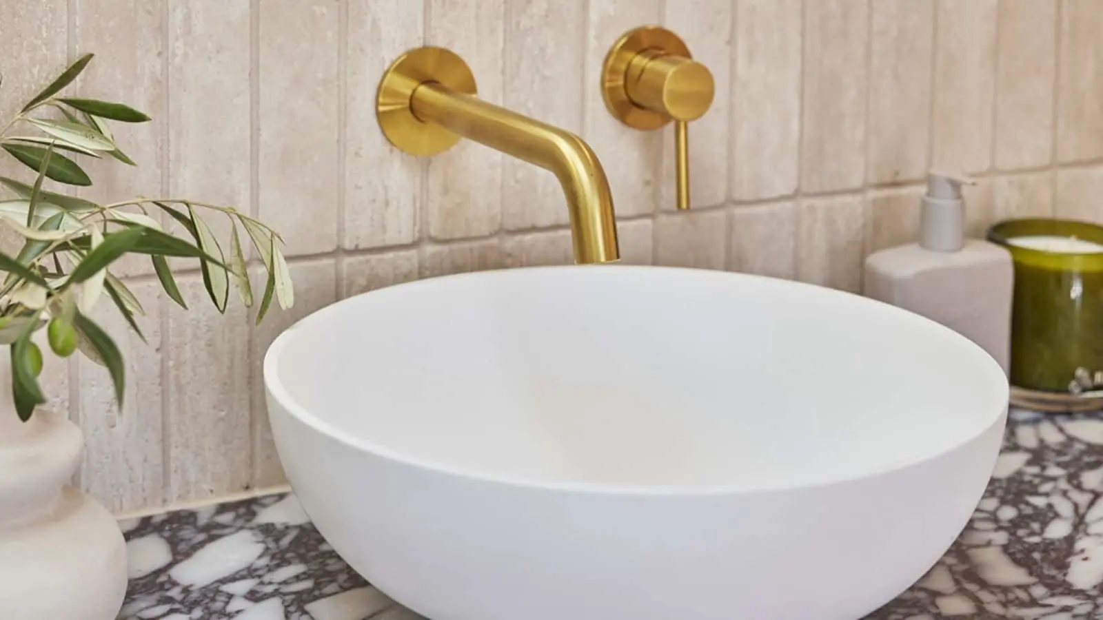 Modern bathroom with white vessel sink, brass wall-mounted faucet, marble countertop, and olive plant decoration.