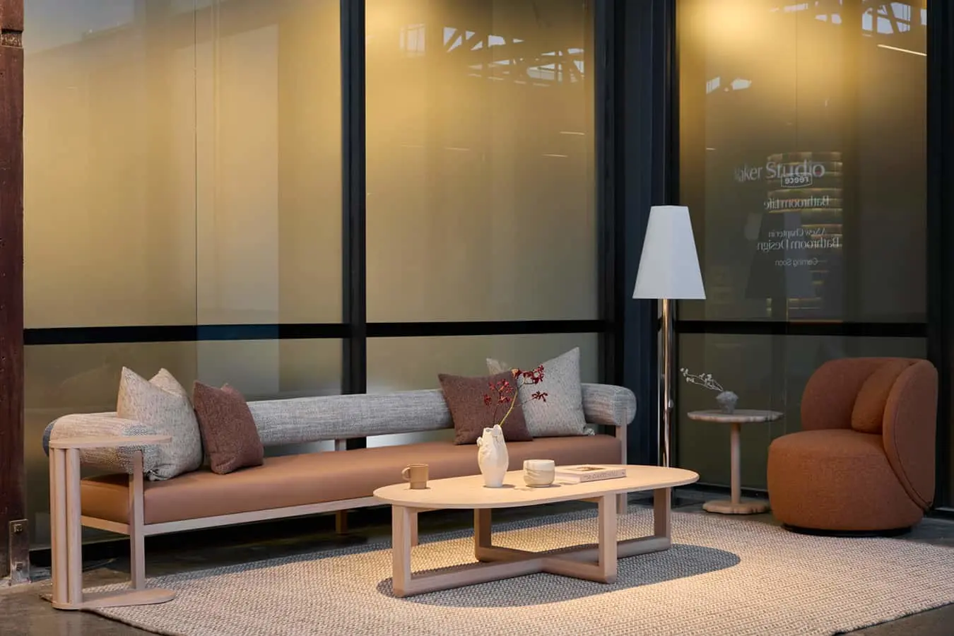 A neutral-toned sofa with cushions, oval coffee table, floor lamp, and rust-colored armchair arranged on a rug near large windows.