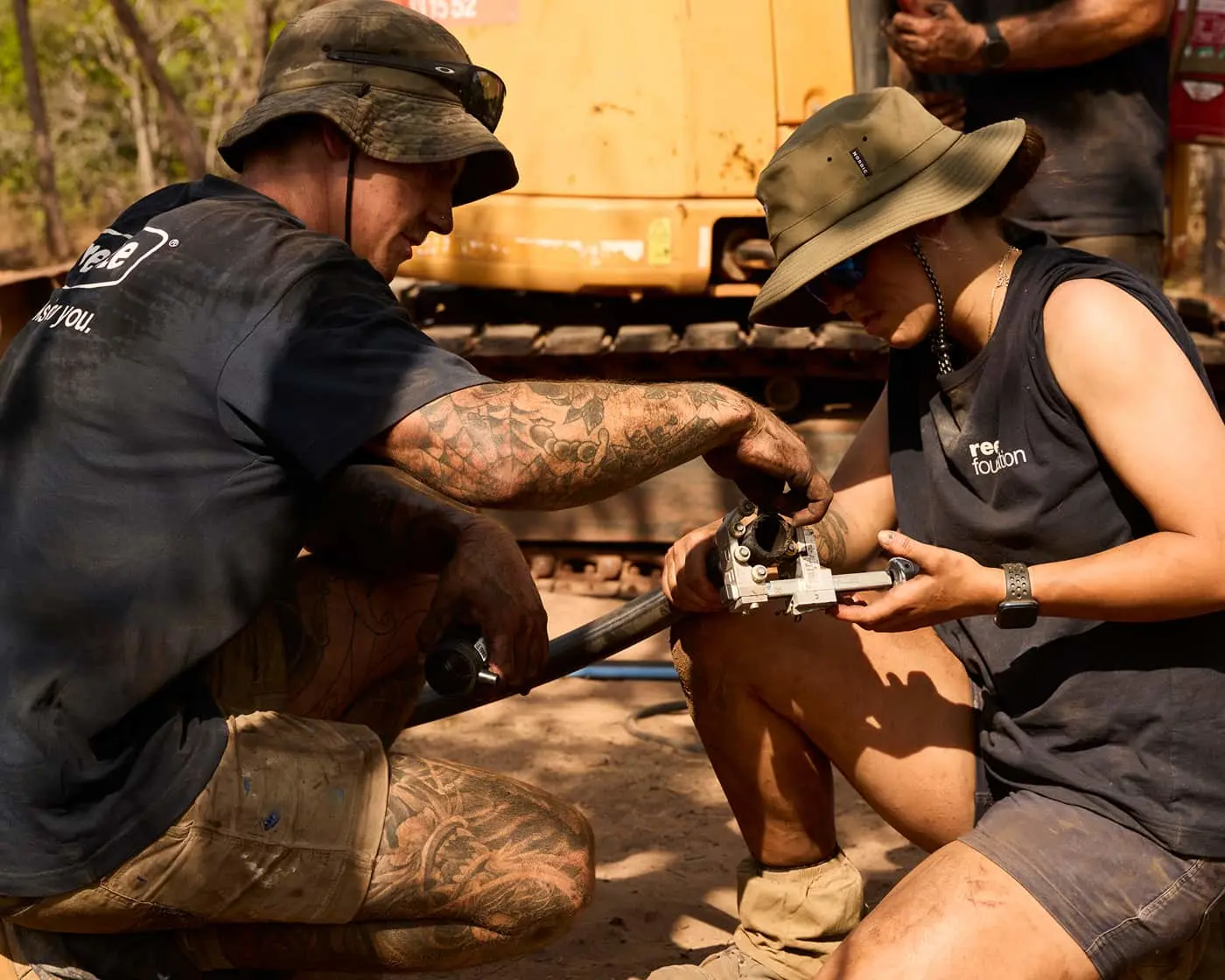 Two people in bucket hats working together on equipment at an outdoor worksite with construction machinery nearby.