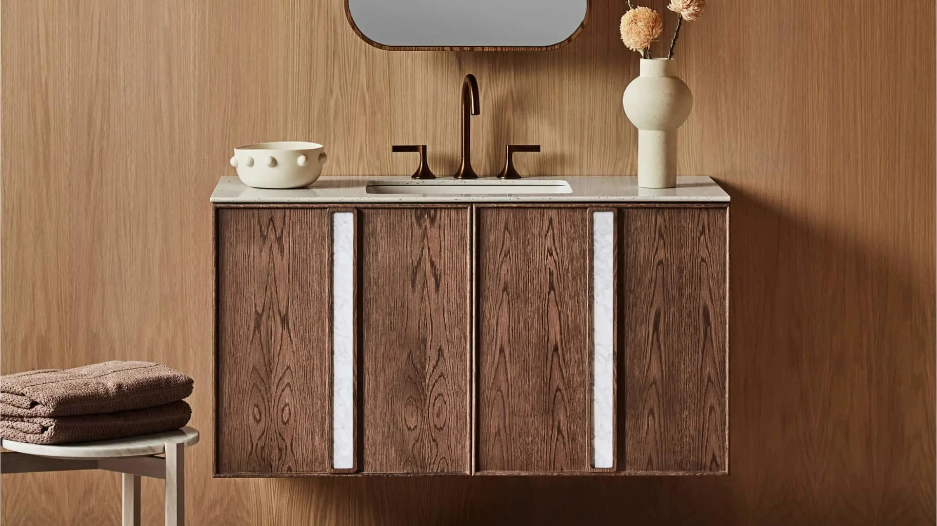 Modern wooden bathroom vanity with marble top, bronze faucet, ceramic bowl and vase, folded towels on side stool.