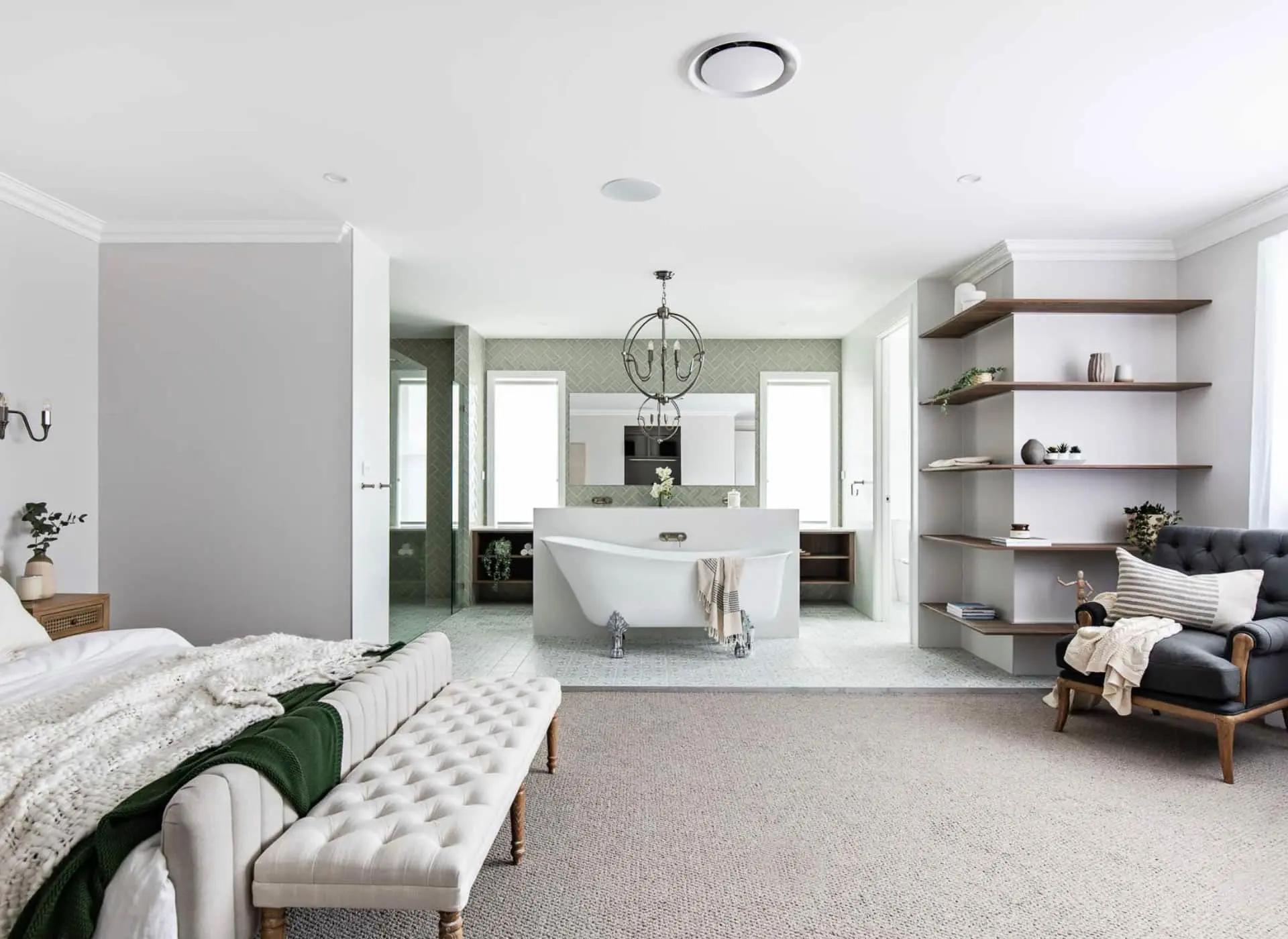Modern bedroom with open bathroom featuring white clawfoot tub, gray armchair, wooden shelves, and neutral color scheme.