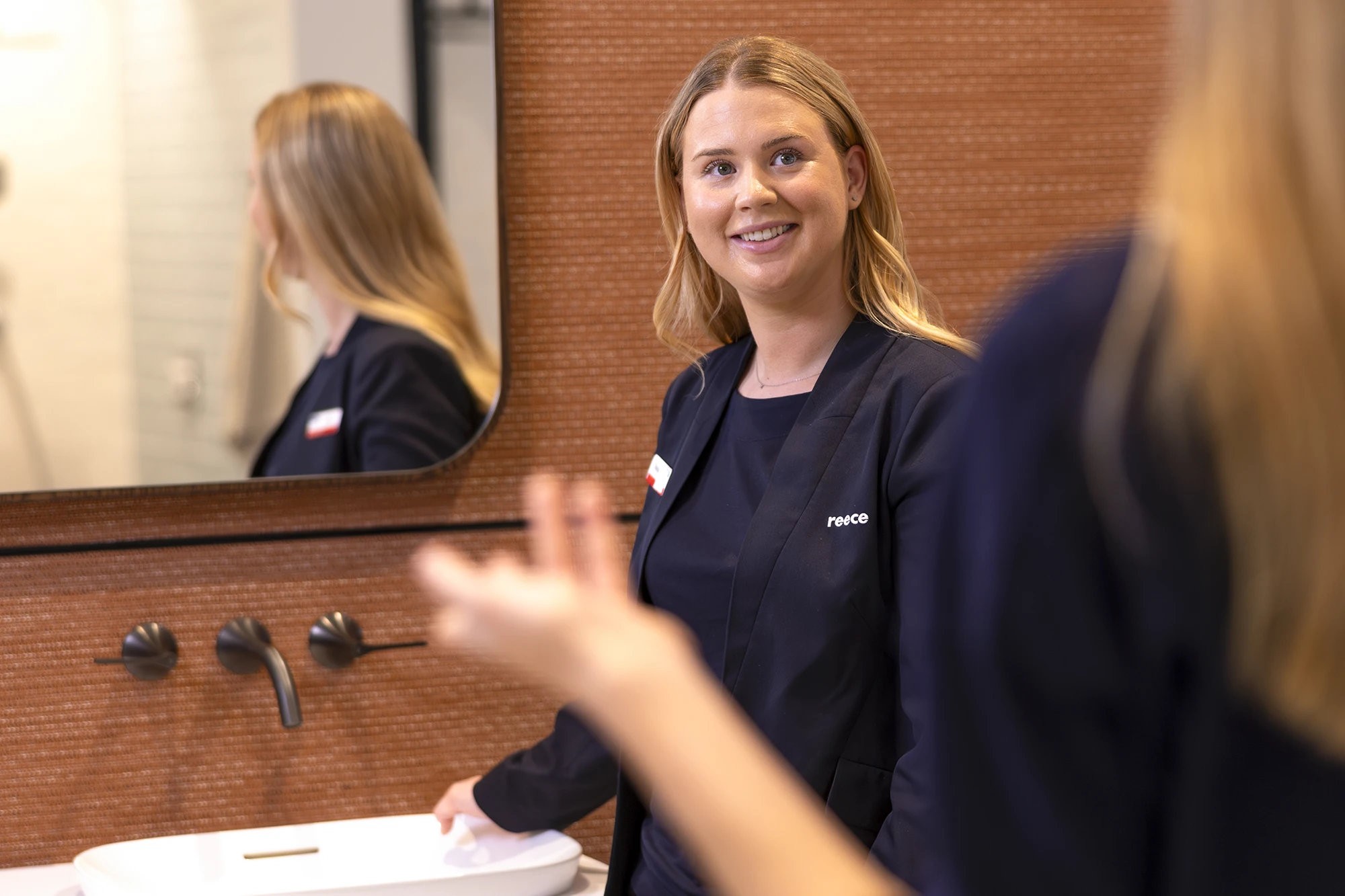 A smiling Reece employee in a navy uniform stands near a bathroom sink display, speaking with a customer.
