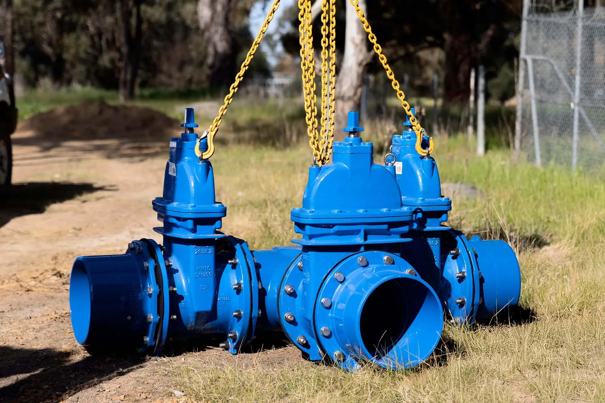 Blue industrial water valves suspended by yellow chains on a dirt path, with trees and a fence in the background.