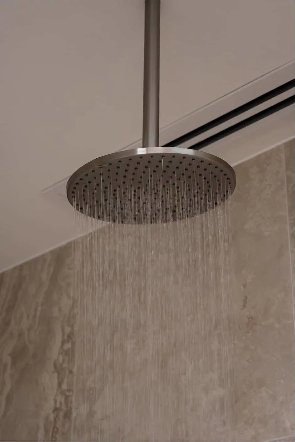 Round ceiling-mounted showerhead with water streaming down in a tiled bathroom.
