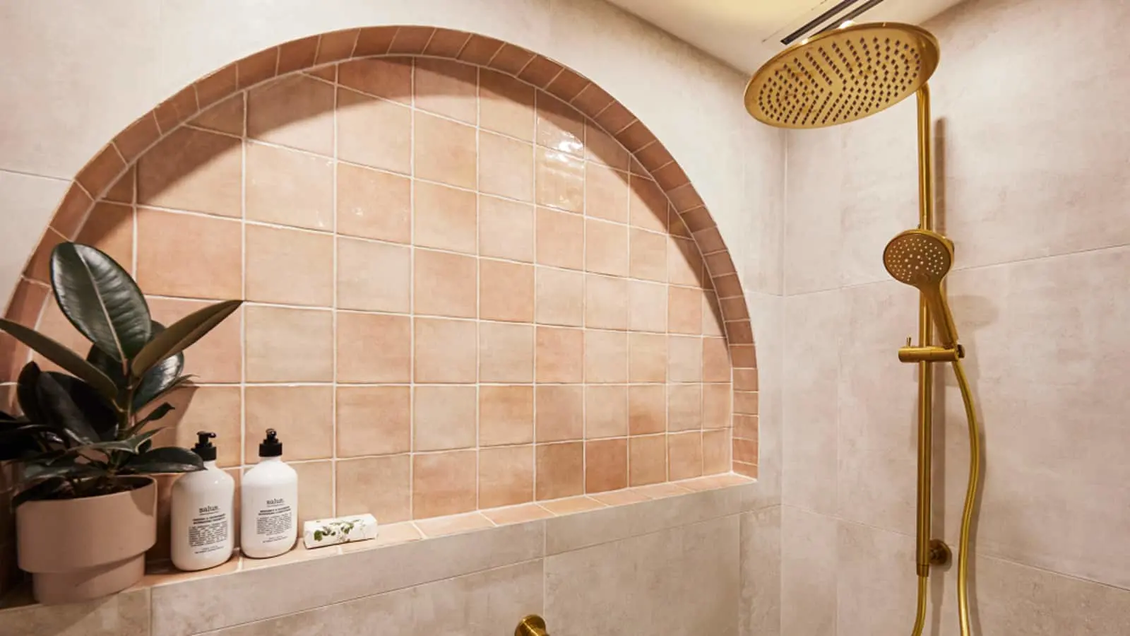 Modern bathroom with terracotta tiled arch niche, gold rainfall shower fixture, and potted plant with white bottles.