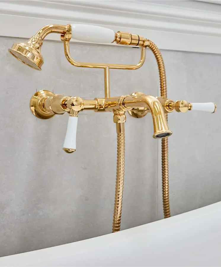 Gold bathtub faucet with handheld shower attachment, featuring white porcelain handles mounted on a gray wall.
