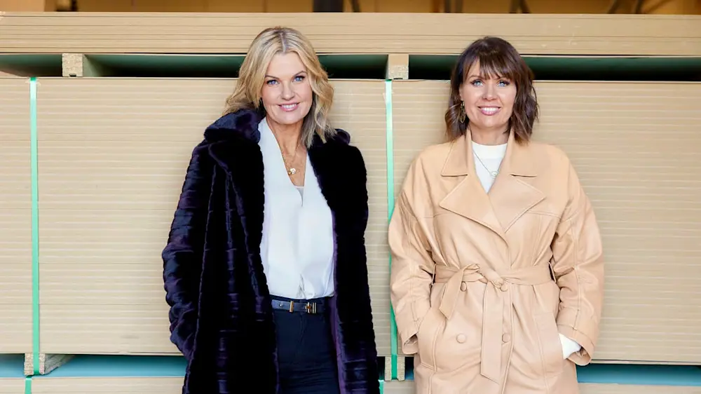 Two women smiling against wooden panels, one wearing a dark coat, the other in a beige trench coat.