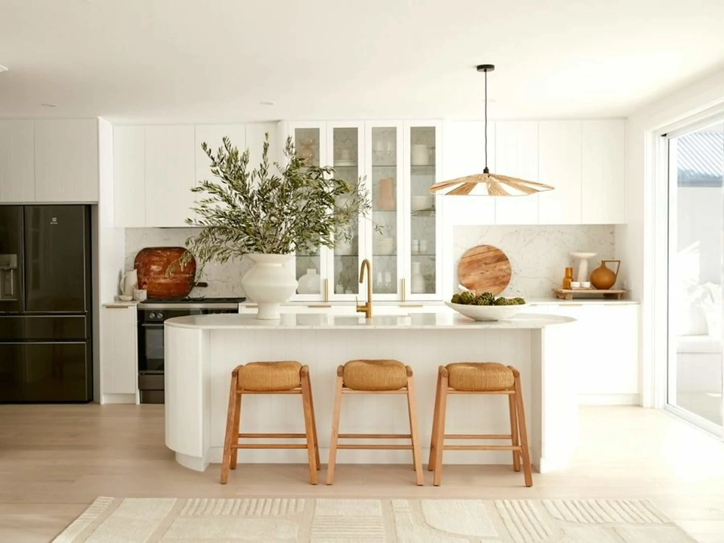 Modern white kitchen with curved island, wooden barstools, pendant light, and olive plant in white vase.