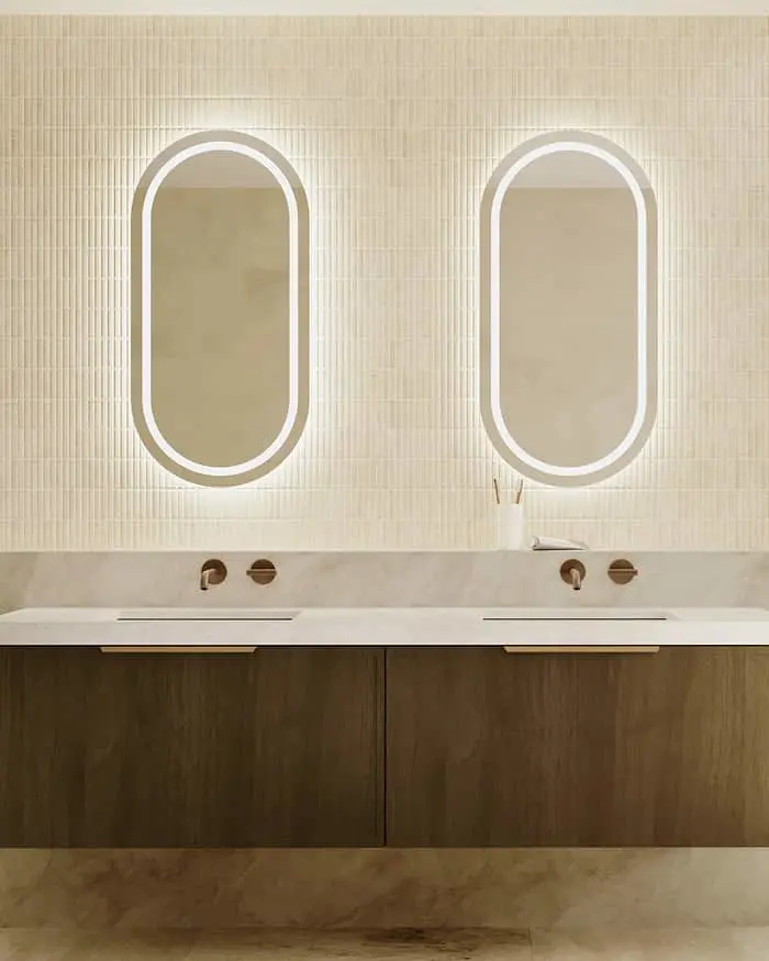 Modern bathroom with two oval backlit mirrors above a double sink vanity with dark wood cabinetry and beige tiled walls.